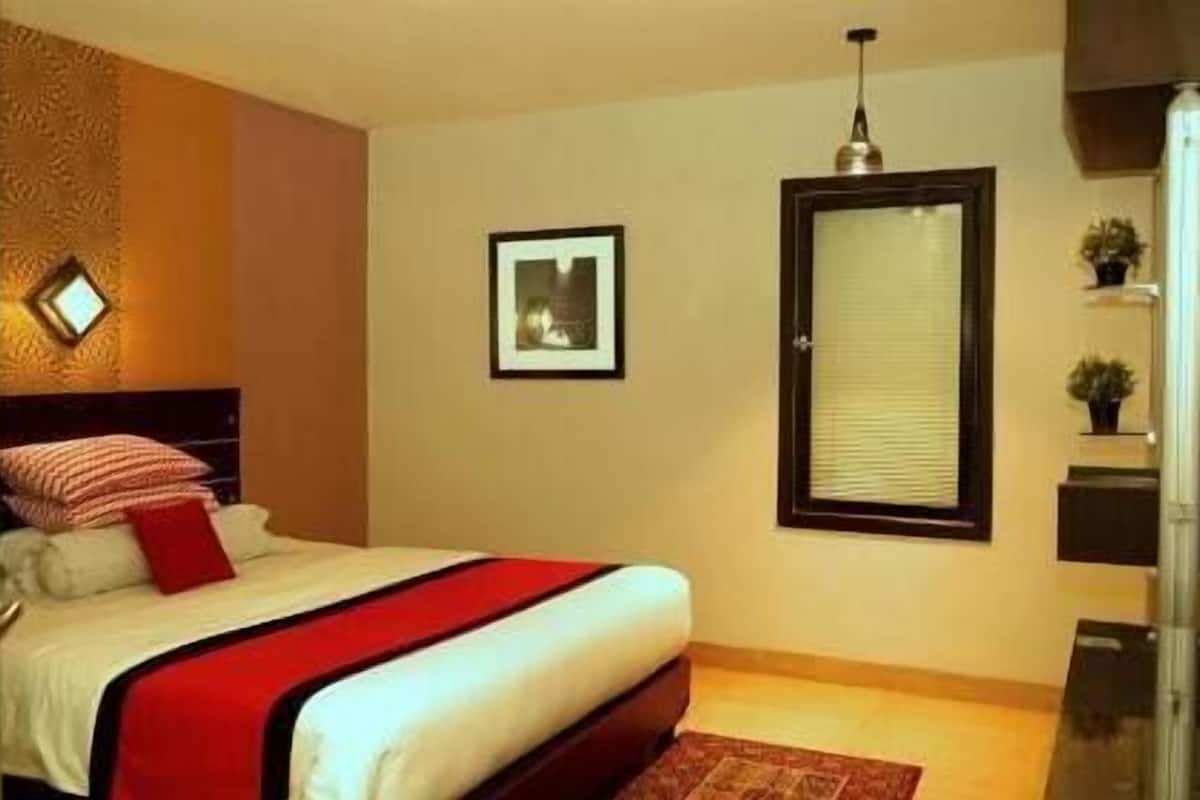 A bedroom with a bed, a framed picture, a window with blinds, and a small plant on a shelf.