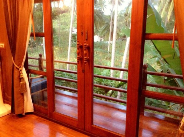 Una habitación con puerta y ventana de madera, piso de madera y vistas a un jardín tropical.