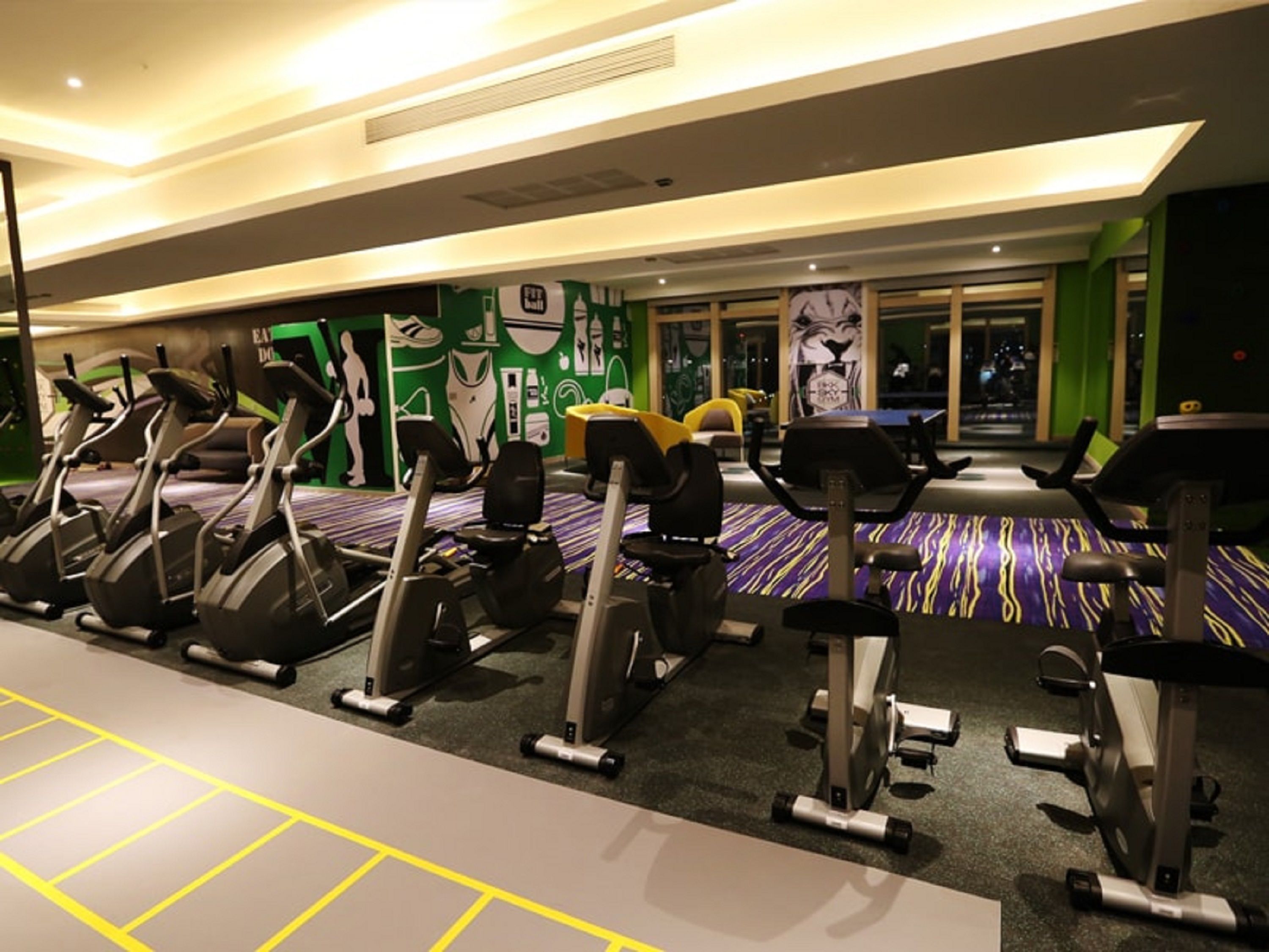 A gym with multiple stationary bikes, a patterned carpet, and green walls with fitness-related graphics.