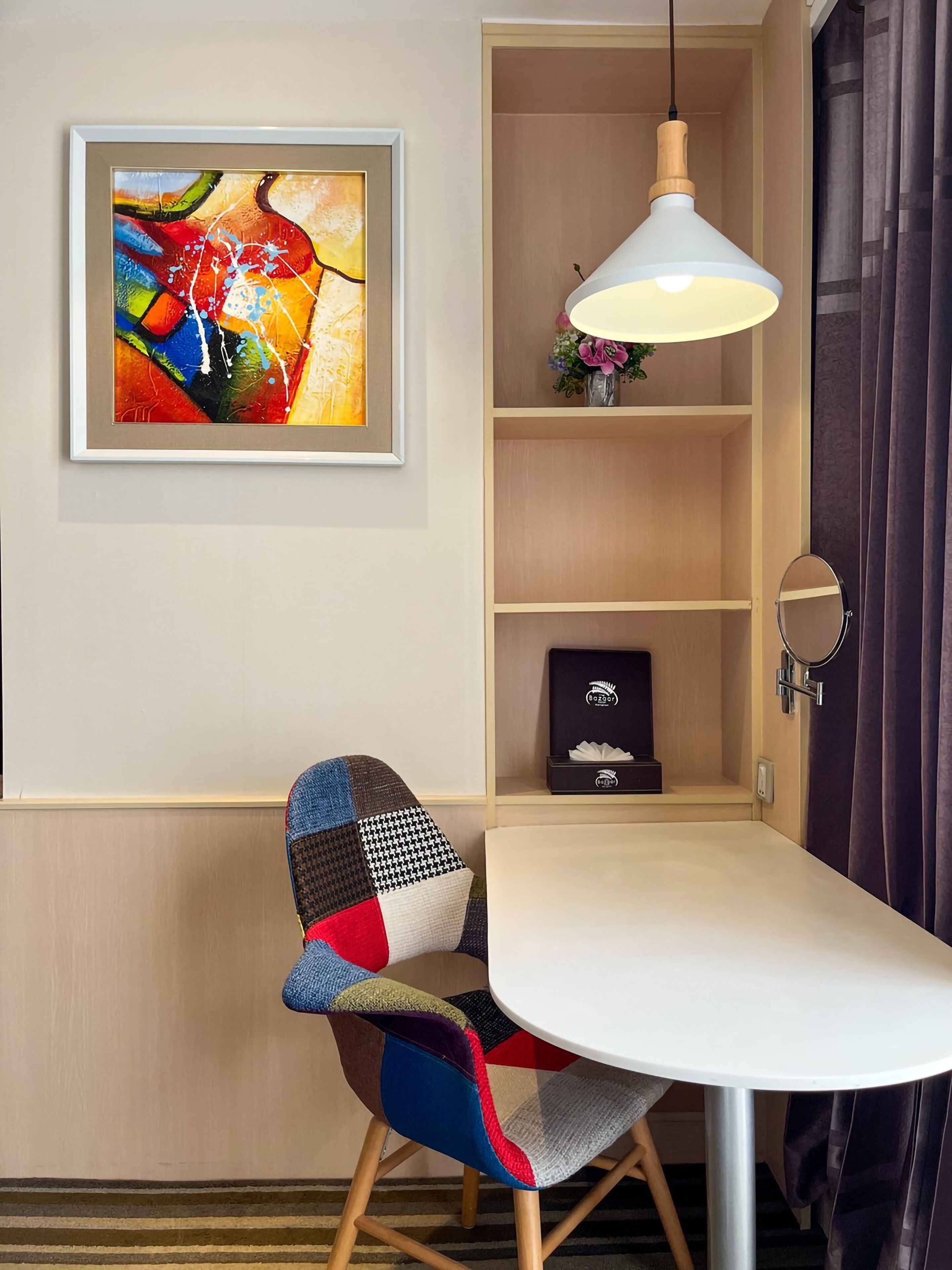 A dining area with a white table, colorful upholstered chairs, a pendant light, a framed abstract painting, and a shelf with a vase and a box.