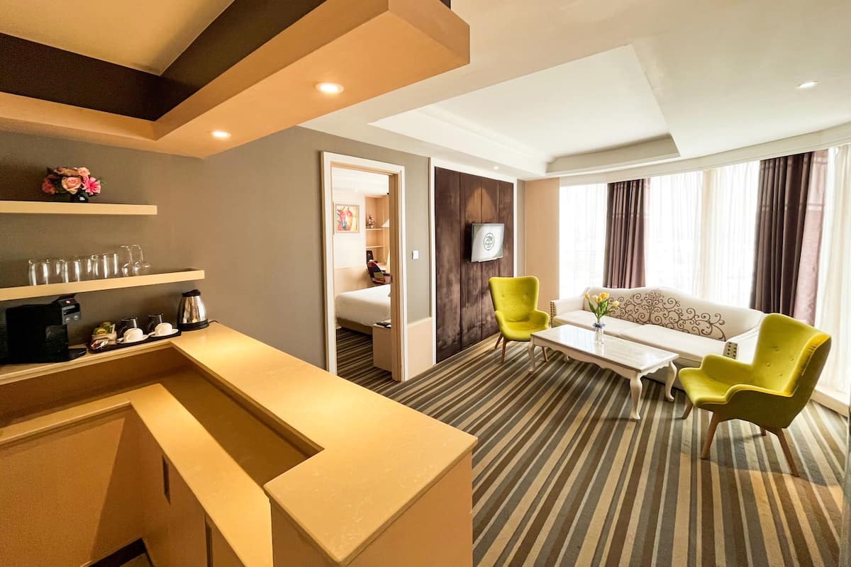 A hotel room with a kitchenette, a sofa, a yellow armchair, a coffee machine, and a TV.