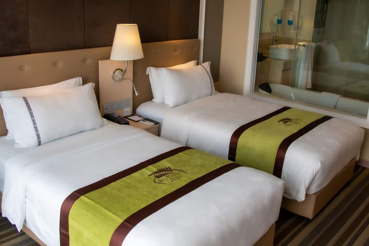 A hotel room with two beds, a nightstand with a lamp, a bathroom visible through a glass partition, and a window with a view.