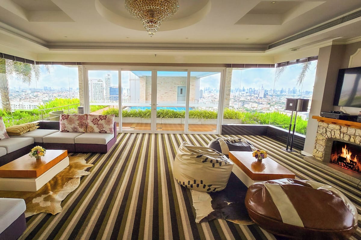 A spacious living room with a large sofa, a fireplace, a television, and a city view through large windows.