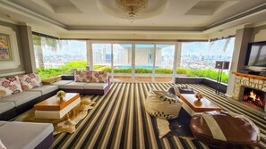 A spacious living room with a large sofa, a fireplace, a television, and a city view through large windows. - The Bazaar Hotel (Bangkok)
