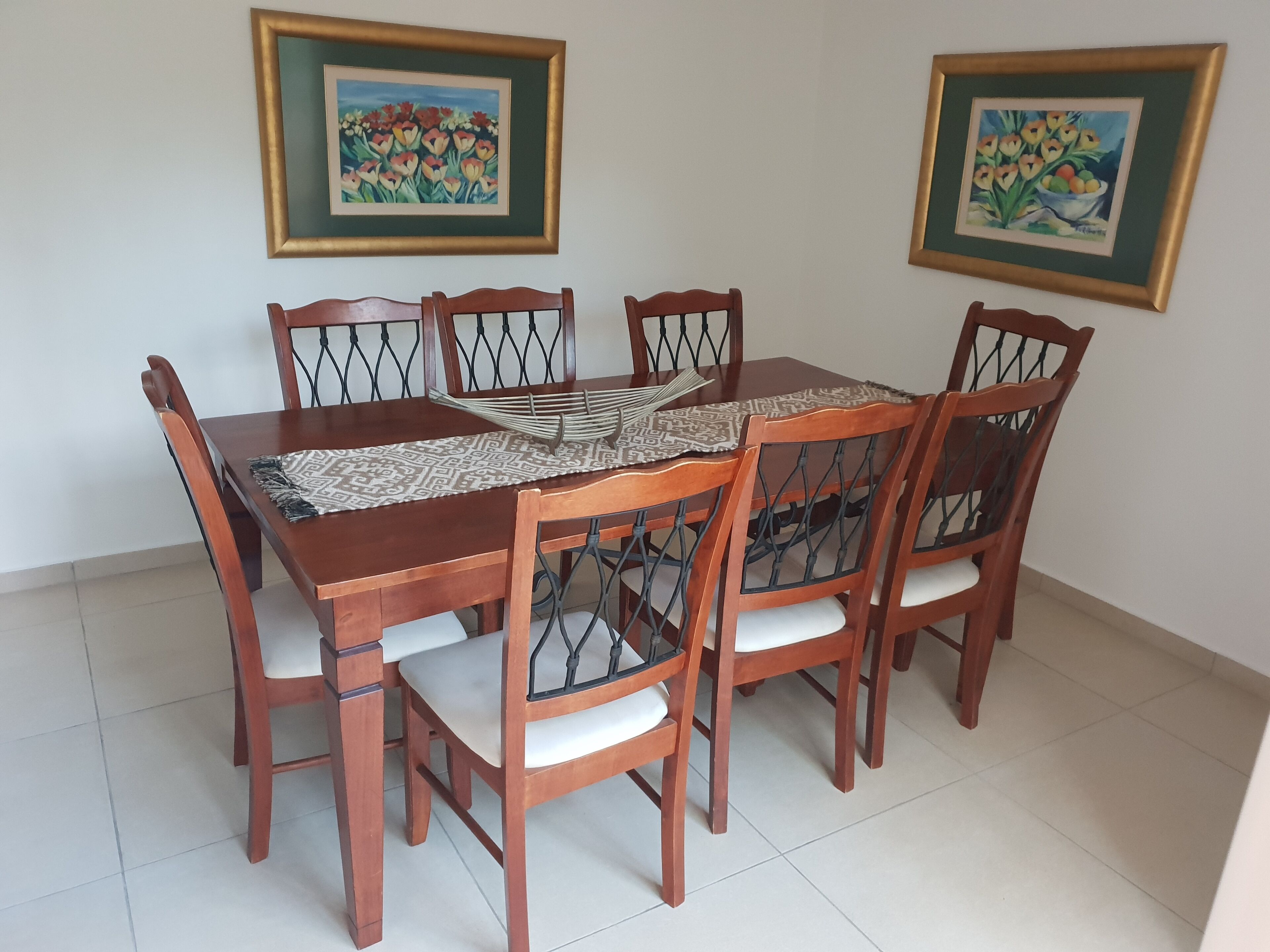 A dining table with six chairs, a patterned table runner, and two framed paintings on the wall.