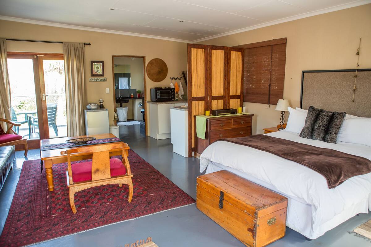 Ein Hotelzimmer mit Bett, einer hölzernen Truhe, einem Schreibtisch mit Stuhl, einer kleinen Küchenzeile und einem Balkon mit Sofa und Tisch.