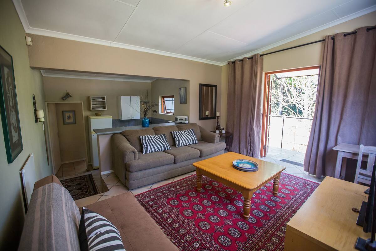 Ein Wohnzimmer mit einer Couch, einem Couchtisch und einem roten Teppich mit Muster. Im Hintergrund ist eine Küchenzeile mit einem Fenster und Vorhängen zu sehen.