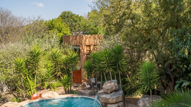 A swimming pool surrounded by rocks and greenery, with a building in the background. - Moonflower Cottages (Randburg)