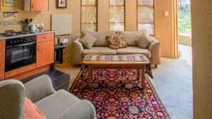 A cozy living room with a sofa, coffee table, and patterned rug. There is a kitchen area with cabinets and a stove. Large windows allow natural light to enter. - Moonflower Cottages (Randburg)