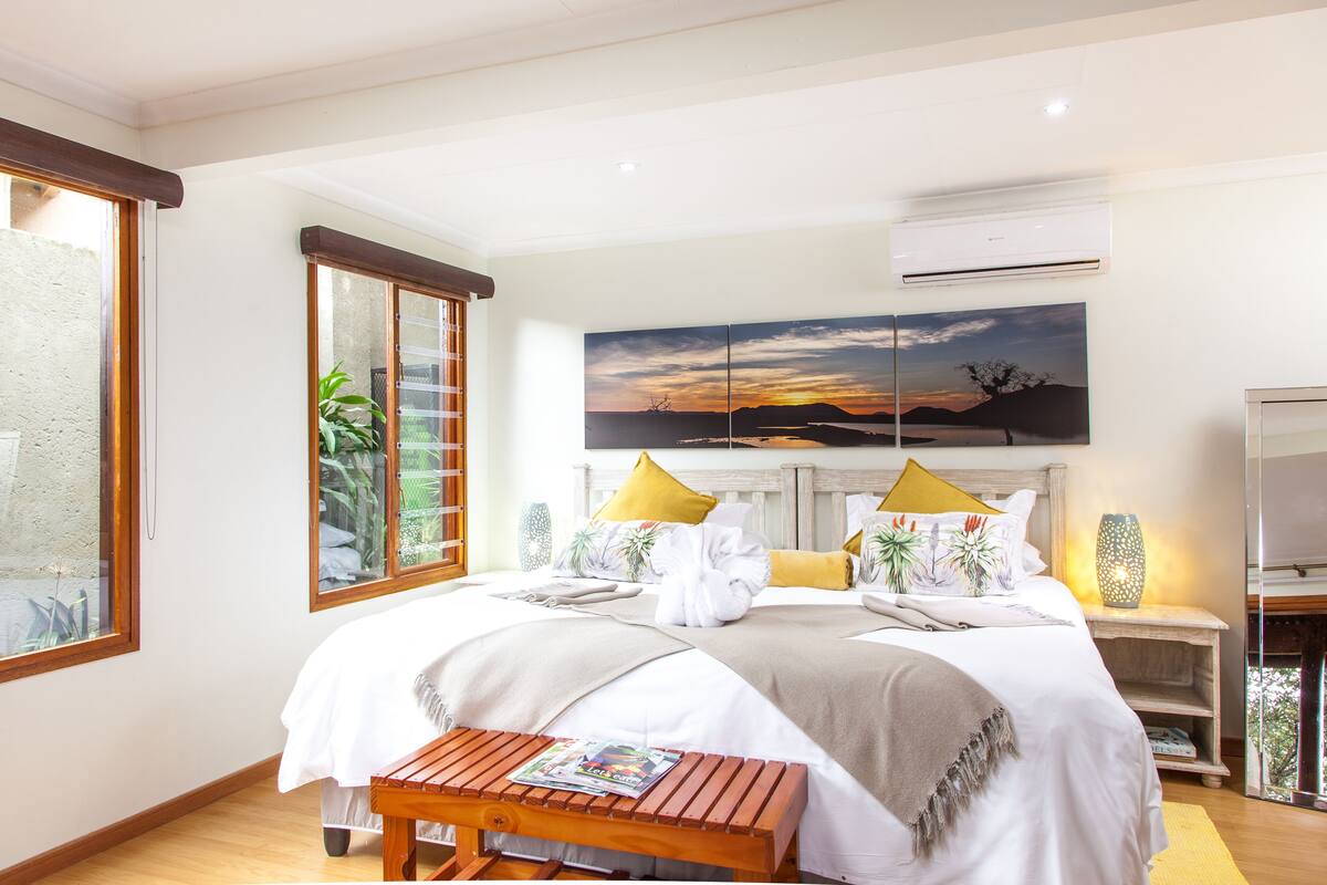 A bedroom with a large bed, a wooden bench, a nightstand, and a wall-mounted air conditioner.