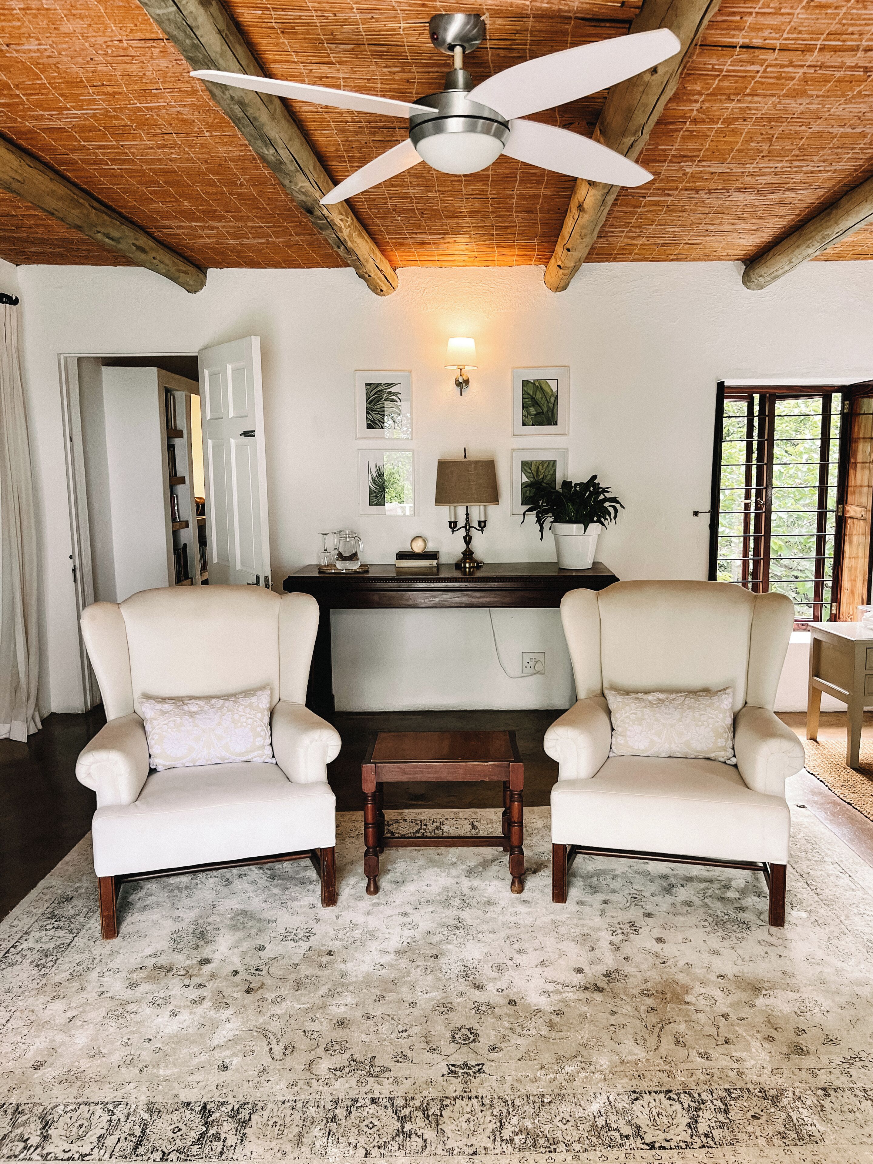 Una habitación con dos sillones, una mesa de madera, un ventilador de techo, una lámpara y cuadros en la pared.