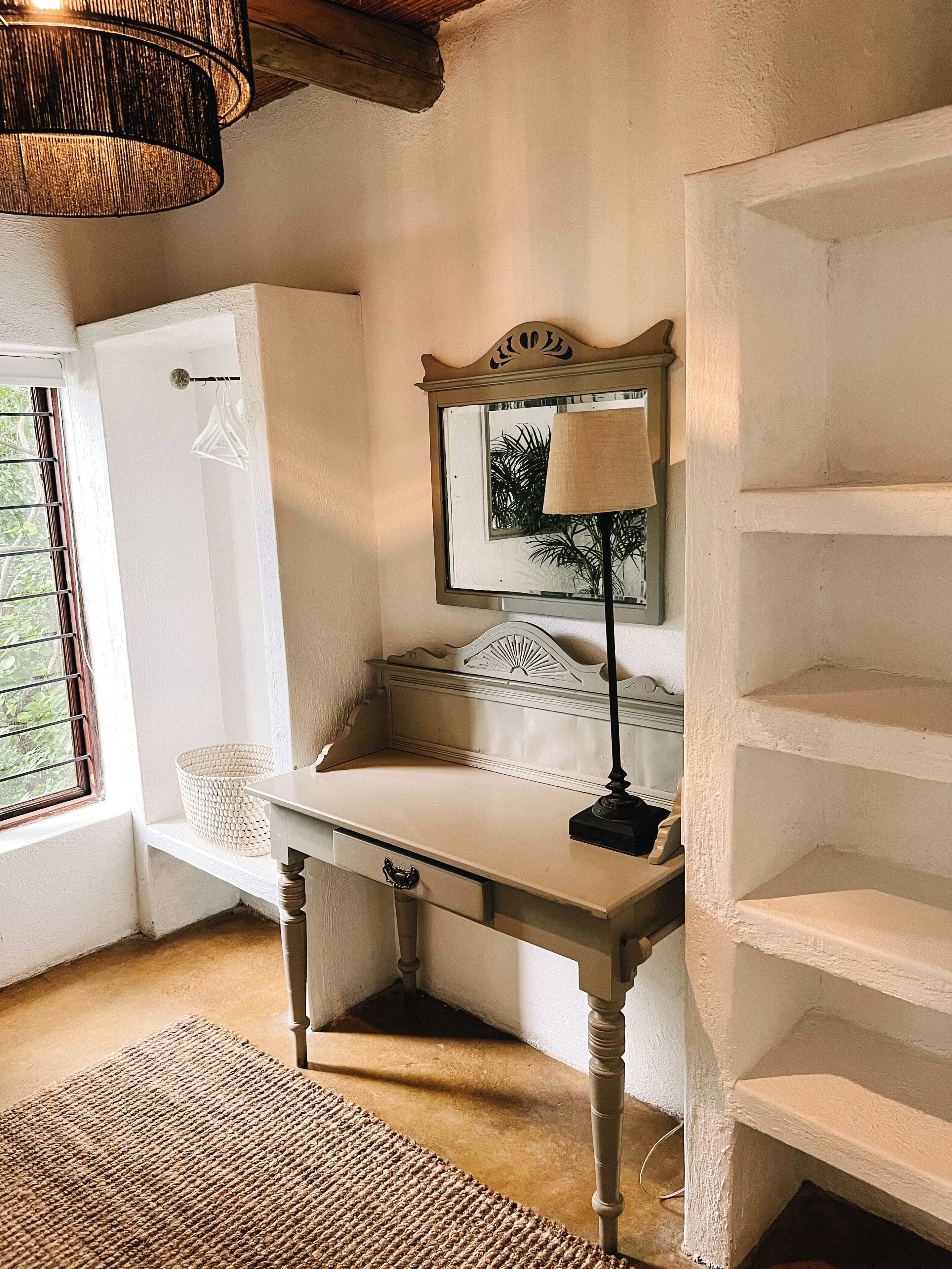 A room with a white desk, a lamp, a mirror, a wicker basket, and a window with blinds.