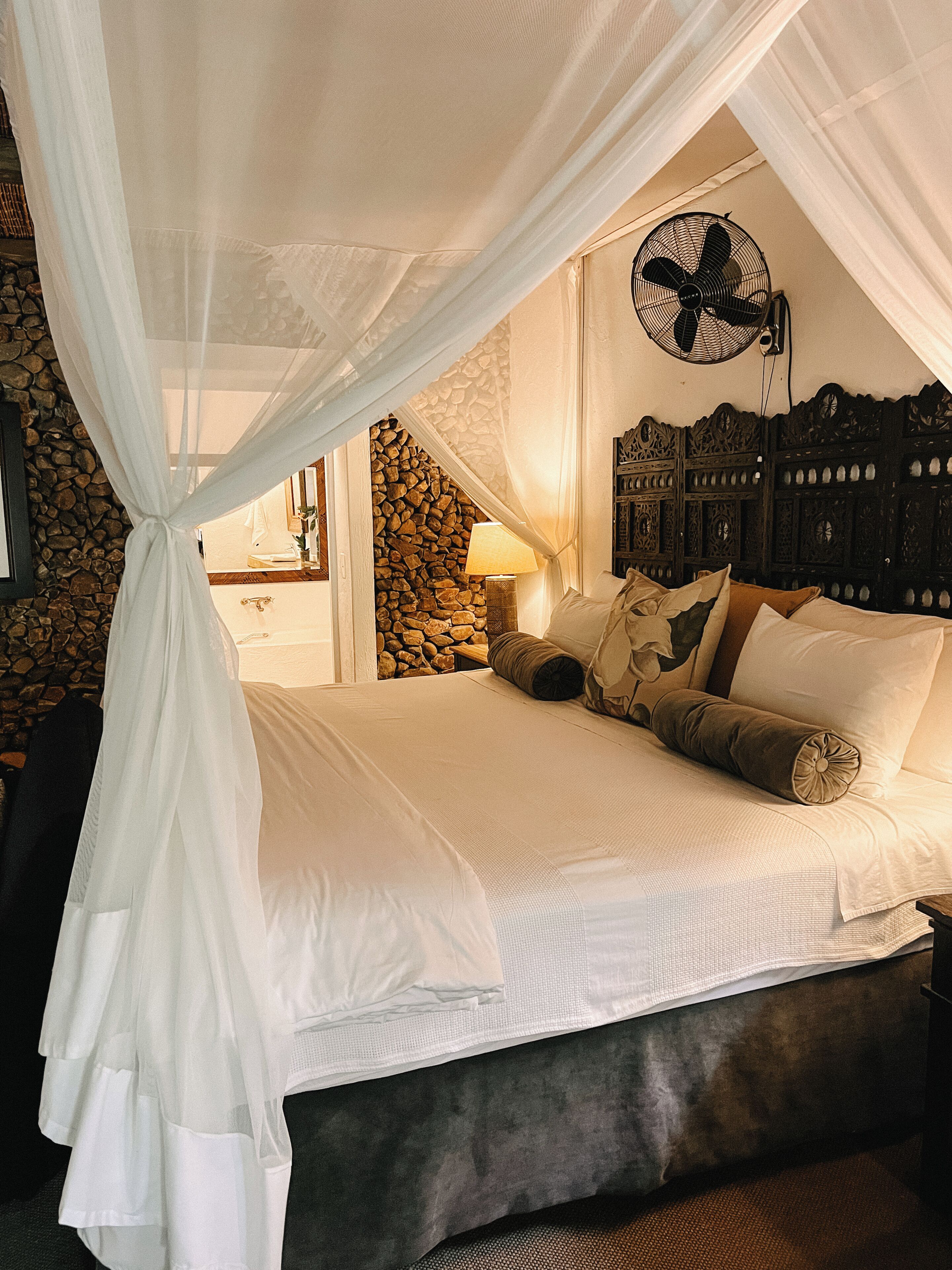 A bed with a canopy, a stone wall, a wooden headboard, and a wall-mounted fan.