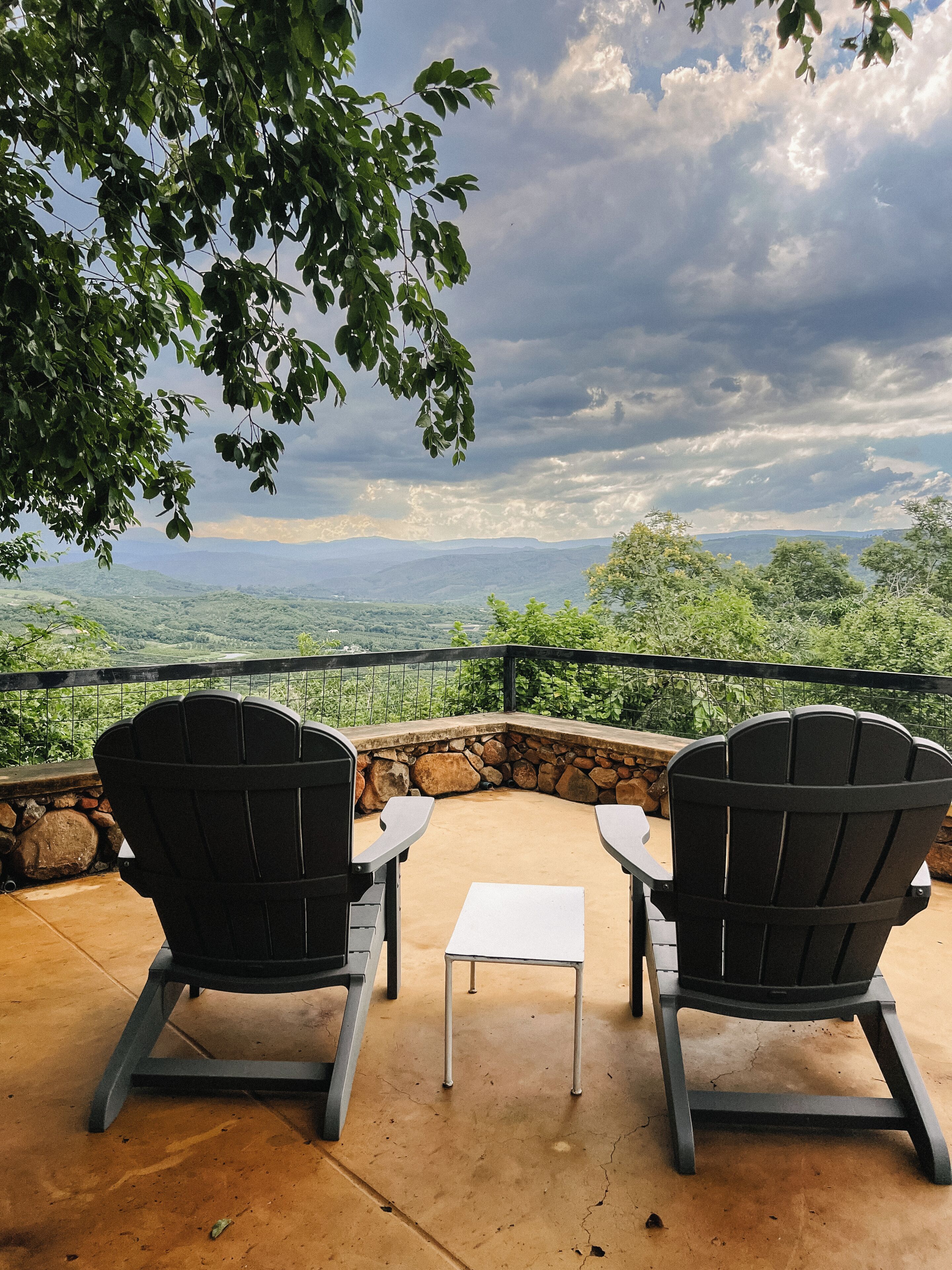 Dos sillas Adirondack mirando un paisaje con un muro de piedra y una pequeña mesa.