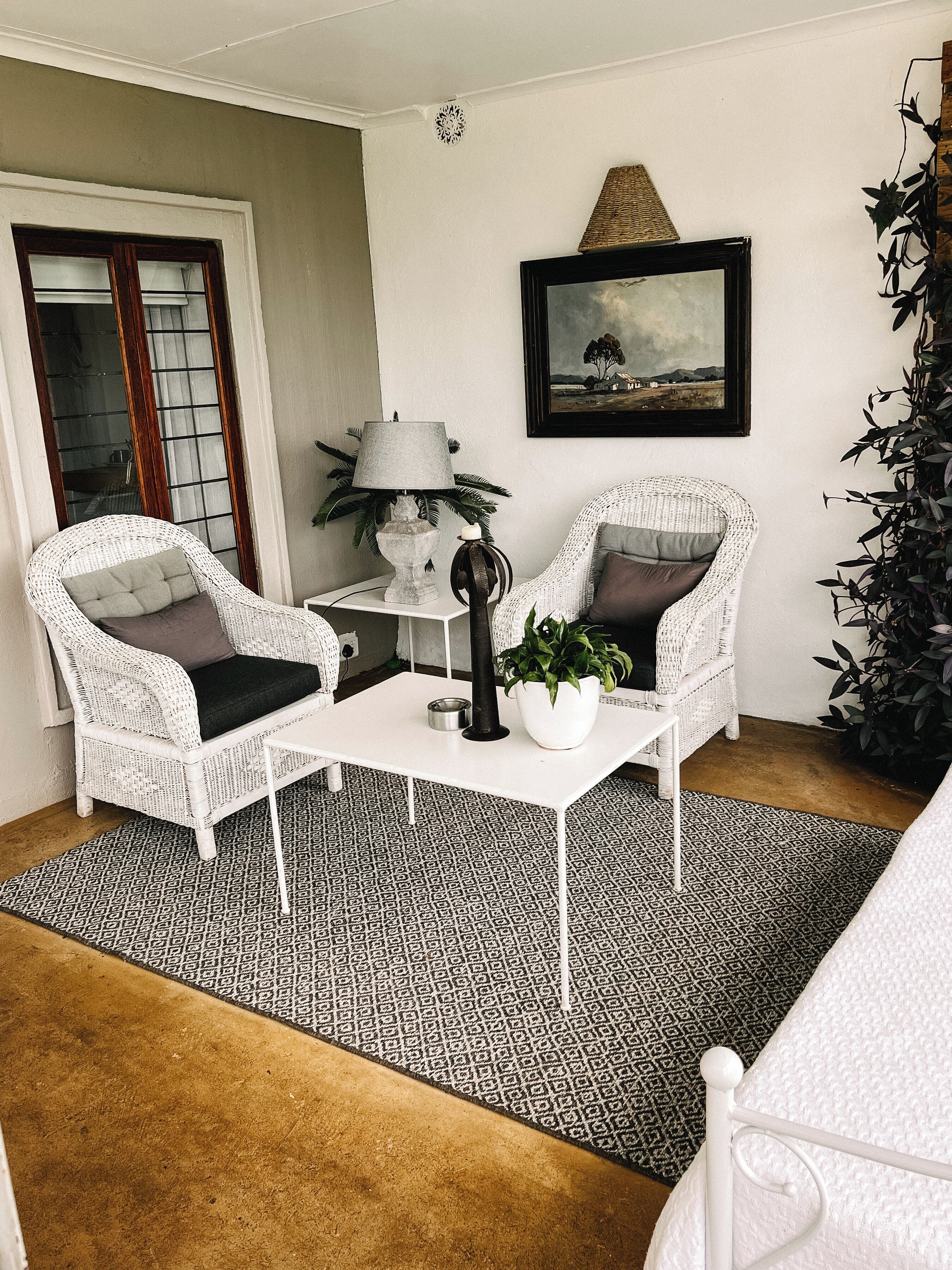 A room with two wicker chairs, a white table, a lamp, a framed picture, and a potted plant.