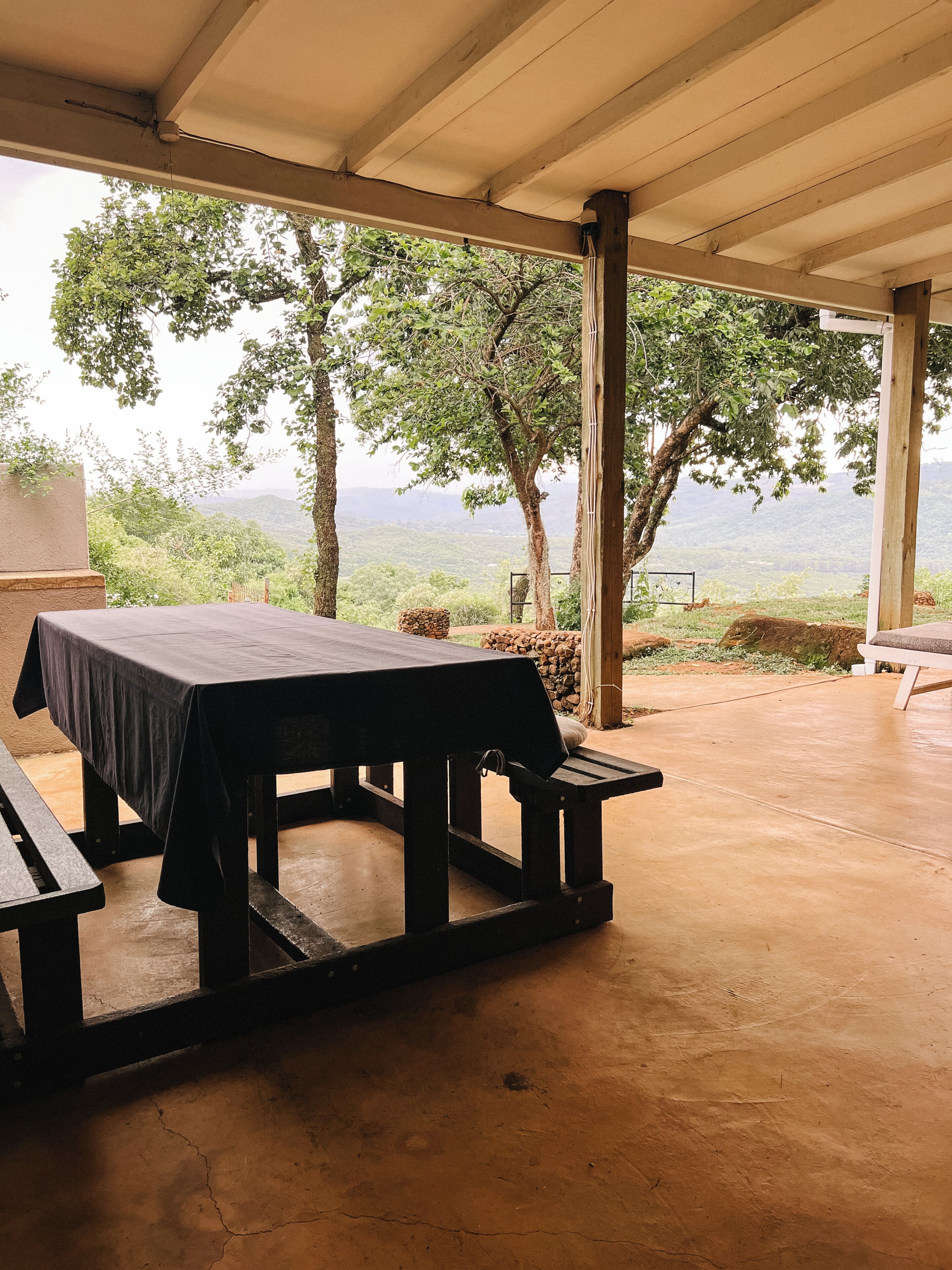 An outdoor covered dining area with a table and benches, surrounded by trees and a view of the landscape.