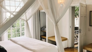 A bedroom with a canopy bed, a bedside table, a nightstand, a window with curtains, and a door leading to another room. - Ashbourne Country Escape (Mbombela)