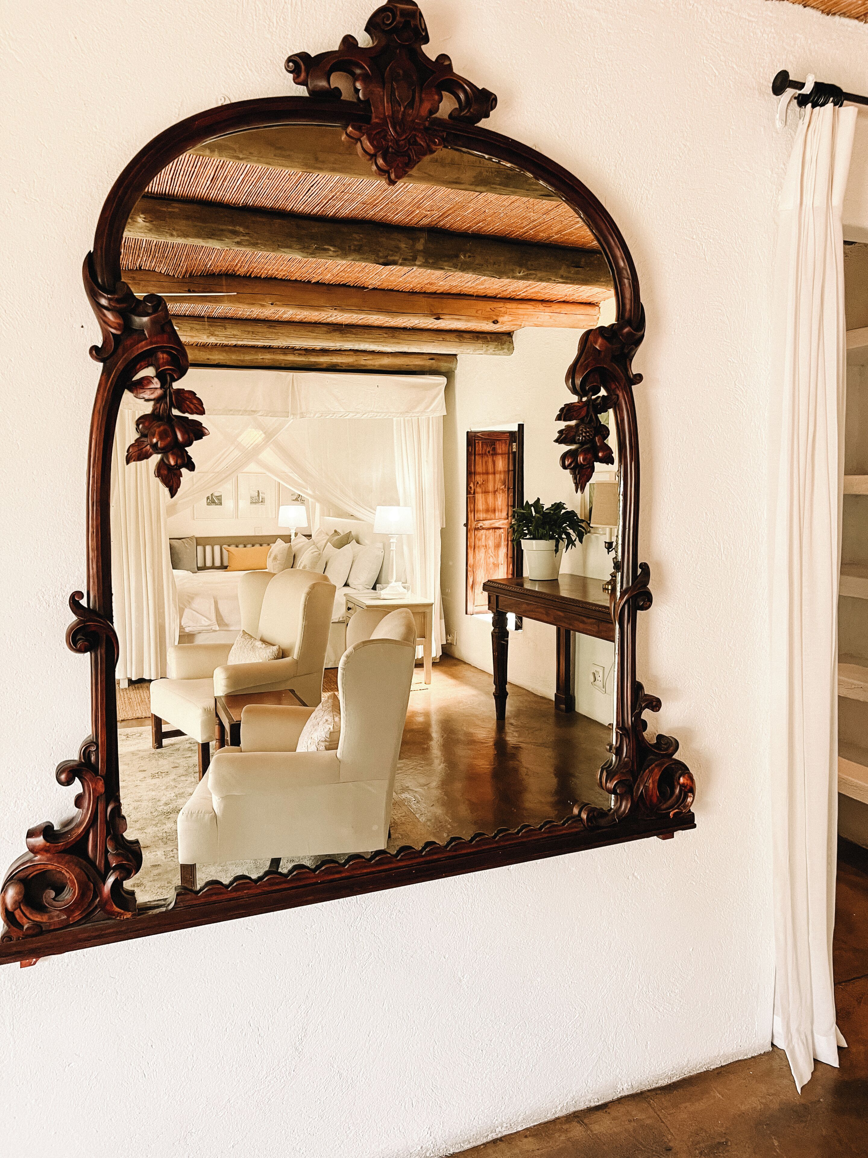 Quarto com um espelho grande e ornamentado, uma cama com roupa de cama branca, uma mesa de madeira e uma cadeira.