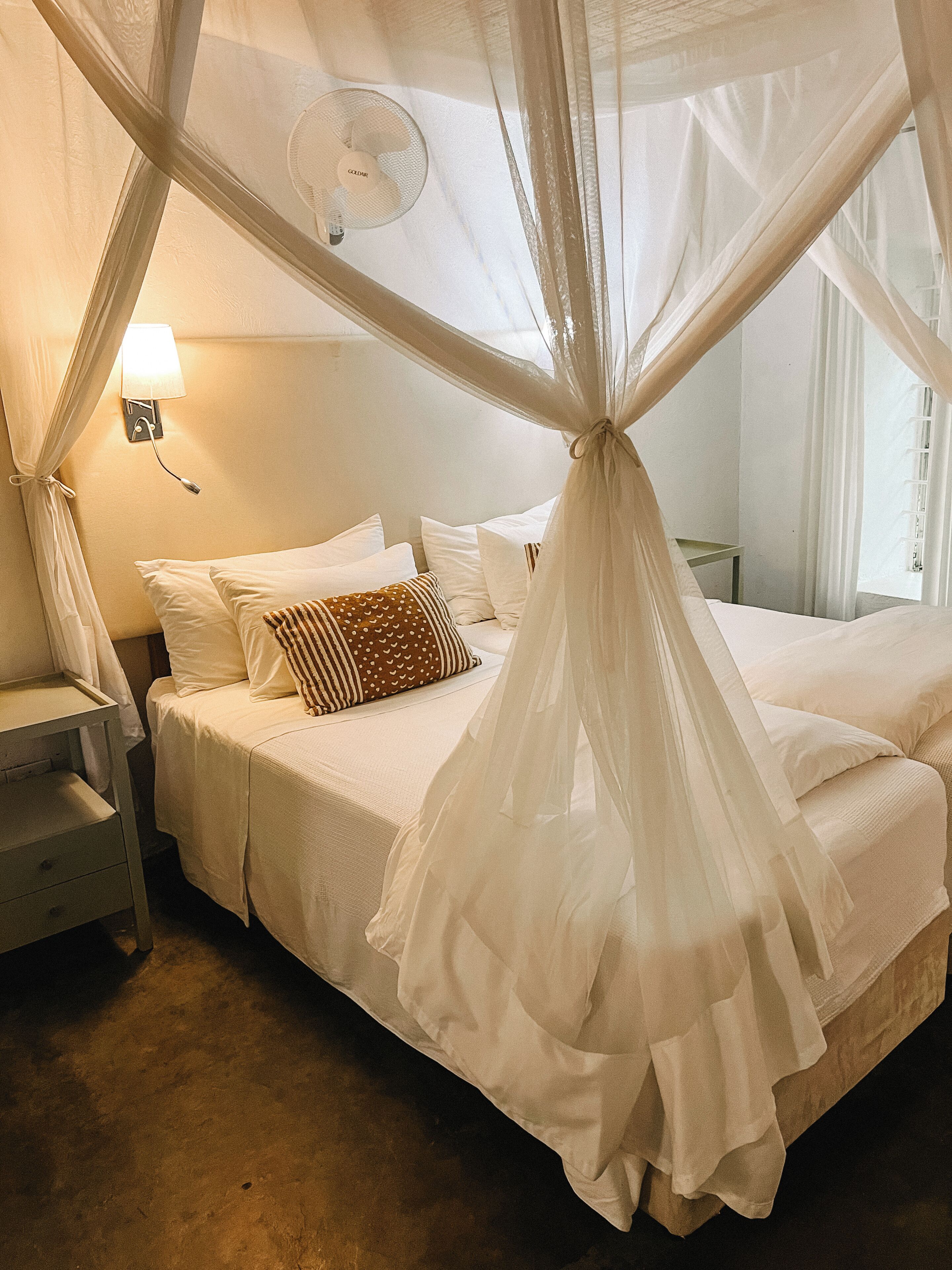 A bed with a canopy, a bedside table, a lamp, and a wall-mounted fan.