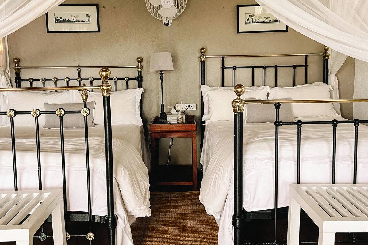Une chambre avec deux lits, une table de chevet avec une lampe, un ventilateur fixé au mur et un tableau encadré accroché au mur.