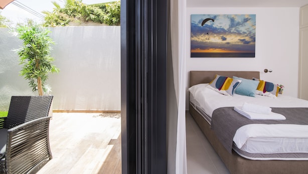 A hotel room with a bed, a window with a view of greenery, a chair, and a painting on the wall. - HaBaitBe Matityahu (Beersheba)
