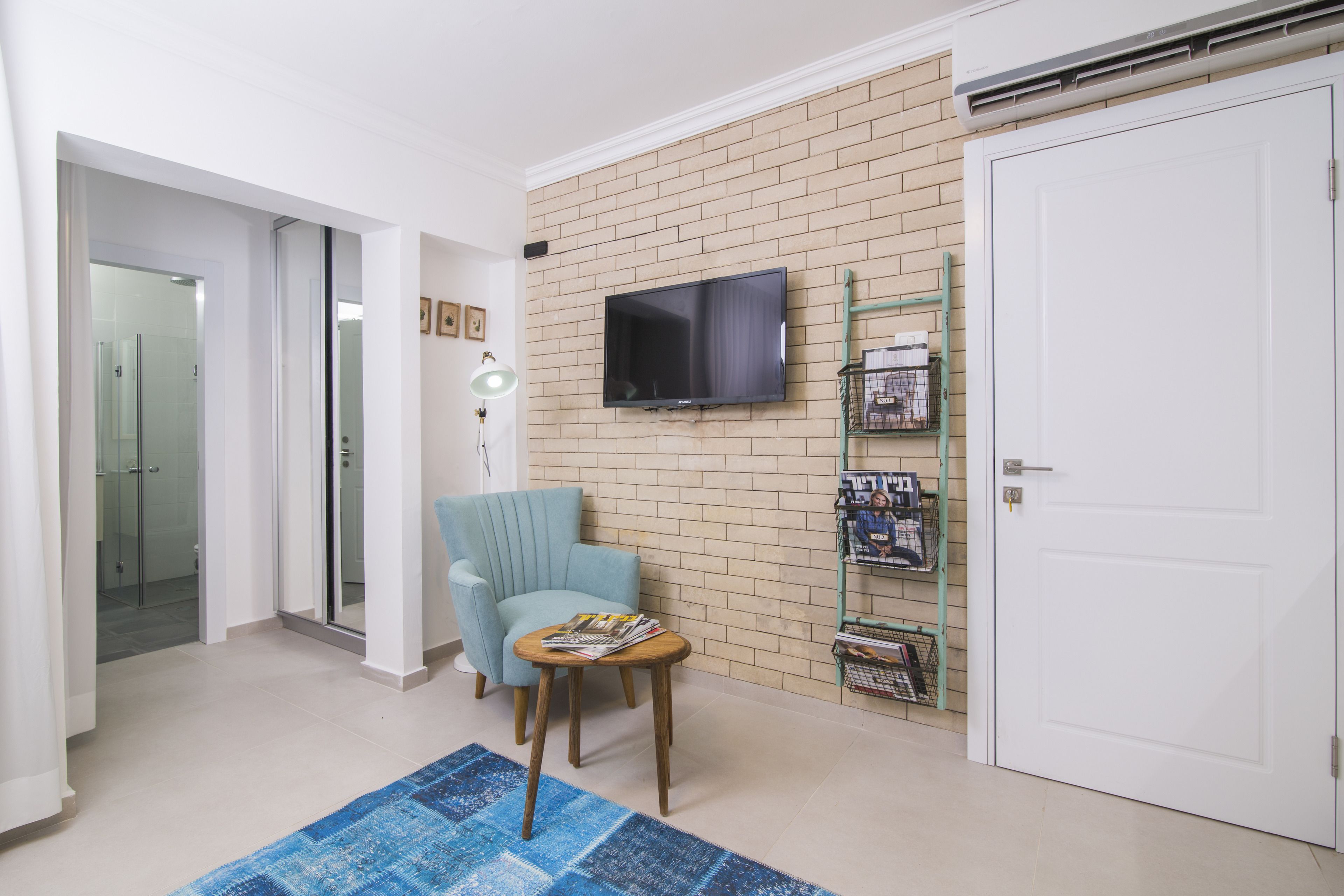 Habitación con pared de ladrillo, televisor montado, sillón azul verdoso, mesita de madera con revistas y una puerta blanca.