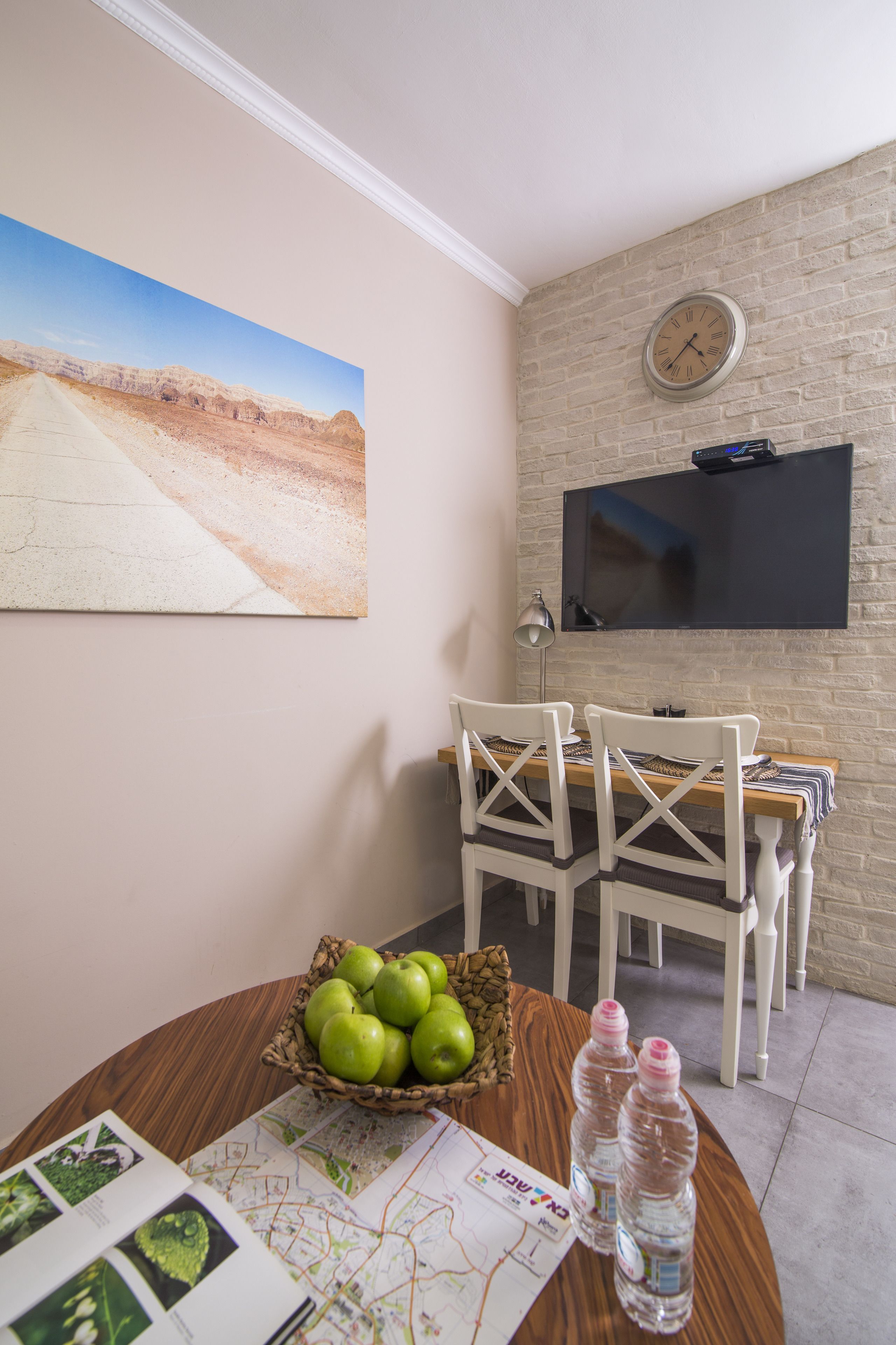 A dining area with a wooden table, chairs, a basket of green apples, water bottles, and a wall-mounted TV.