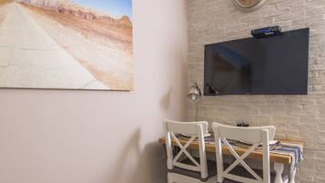 A dining area with a wooden table, chairs, a basket of green apples, water bottles, and a wall-mounted TV.