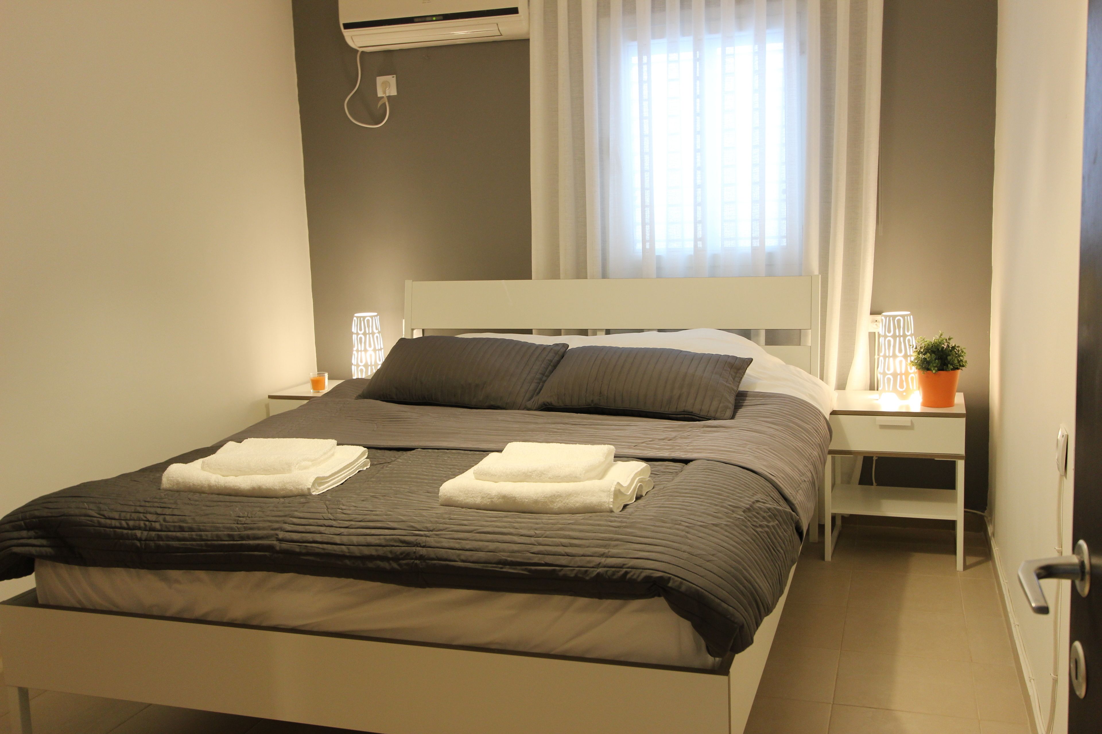 A neatly made bed with two folded towels, a bedside table with a lamp, and a potted plant.