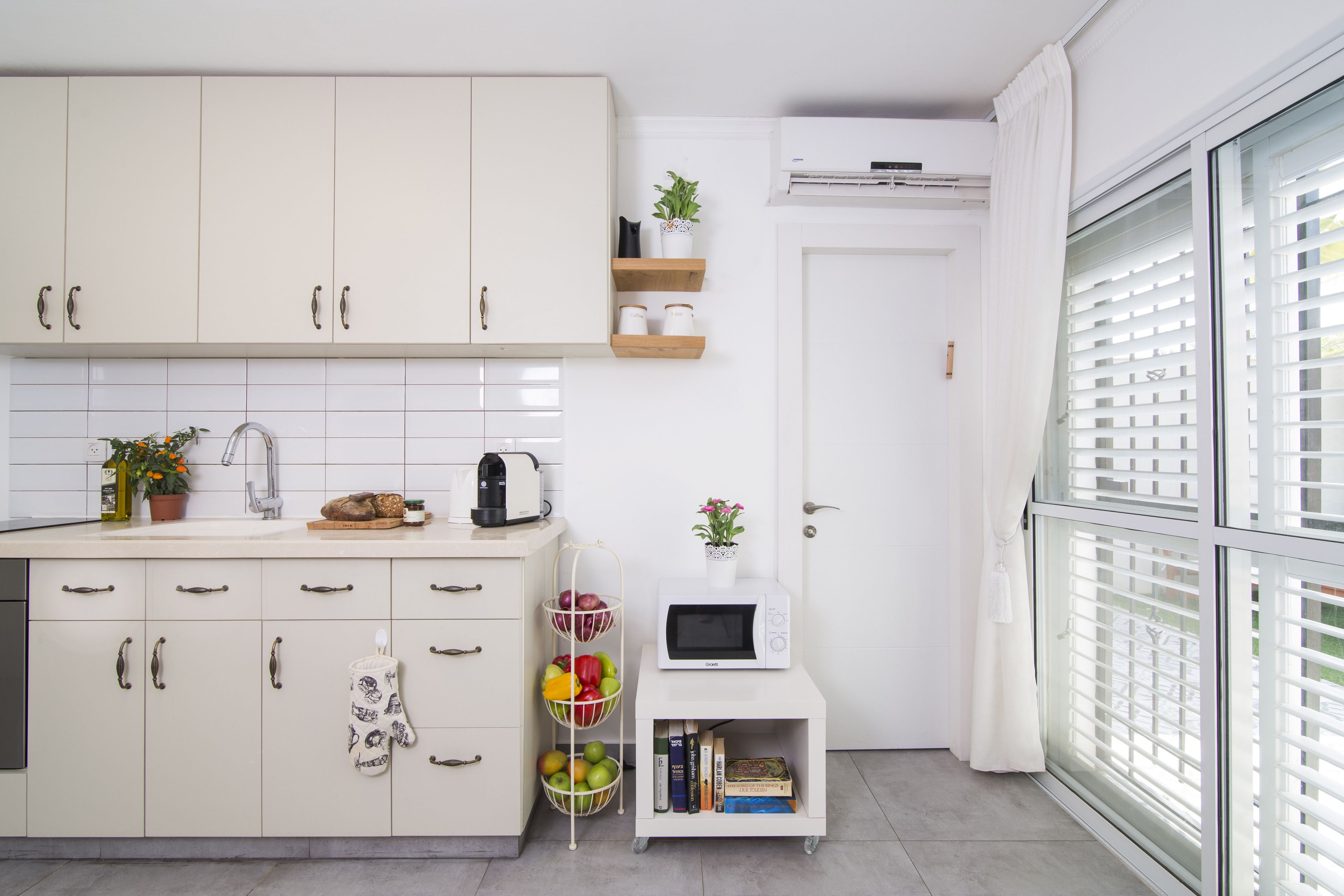 A modern kitchen with white cabinets, a microwave, a small shelf with plants, and a window with blinds.