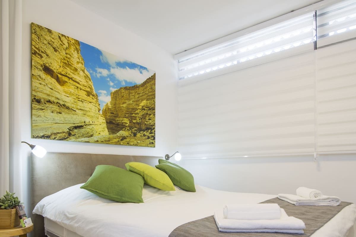 A bedroom with a bed, pillows, a bedside table with a plant, a painting on the wall, and a window with blinds.