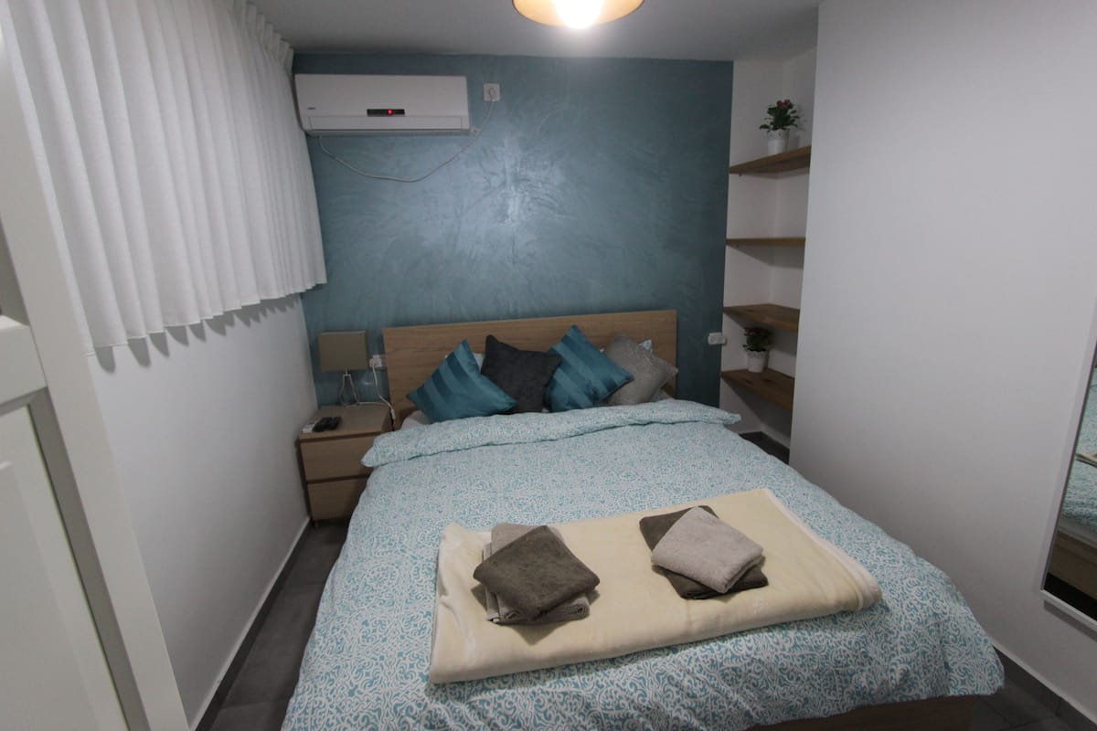 A bedroom with a bed, a nightstand, a wall-mounted air conditioner, and a window with curtains.