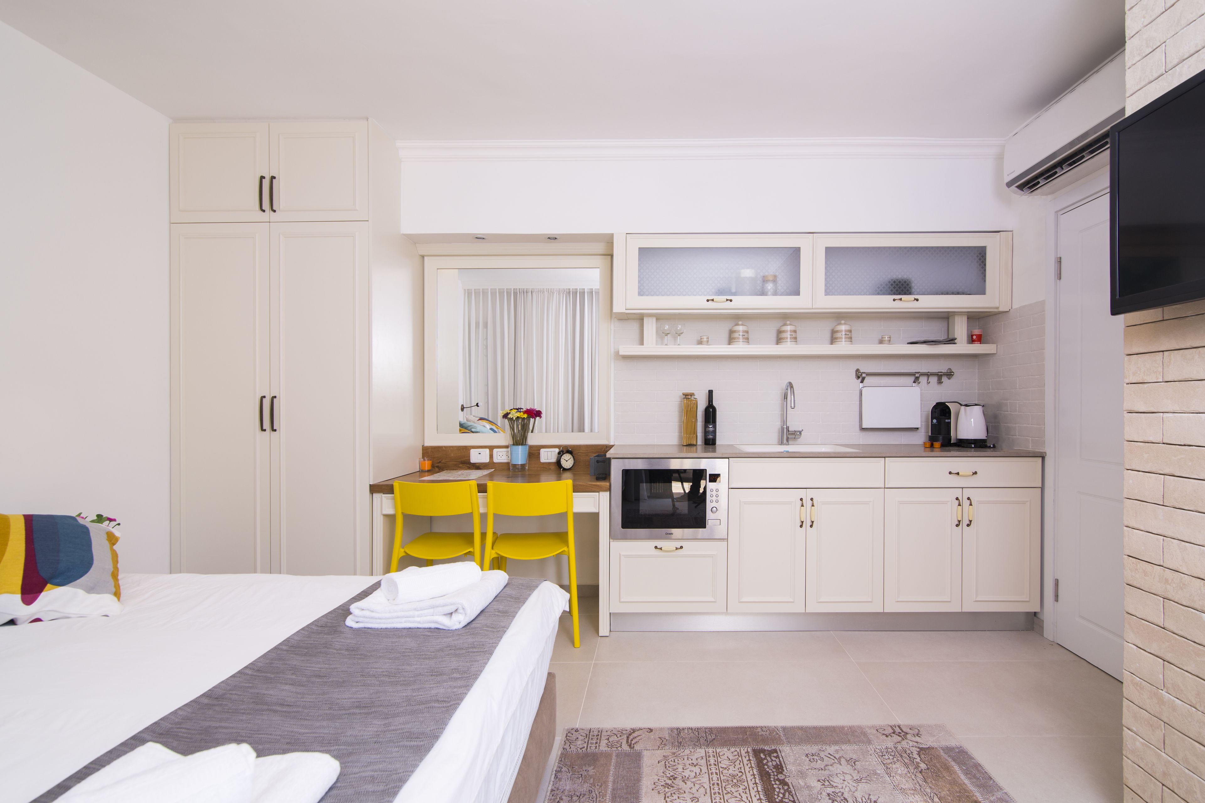 Habitación de hotel con cama, un escritorio pequeño con silla, kitchenette y televisor en la pared.