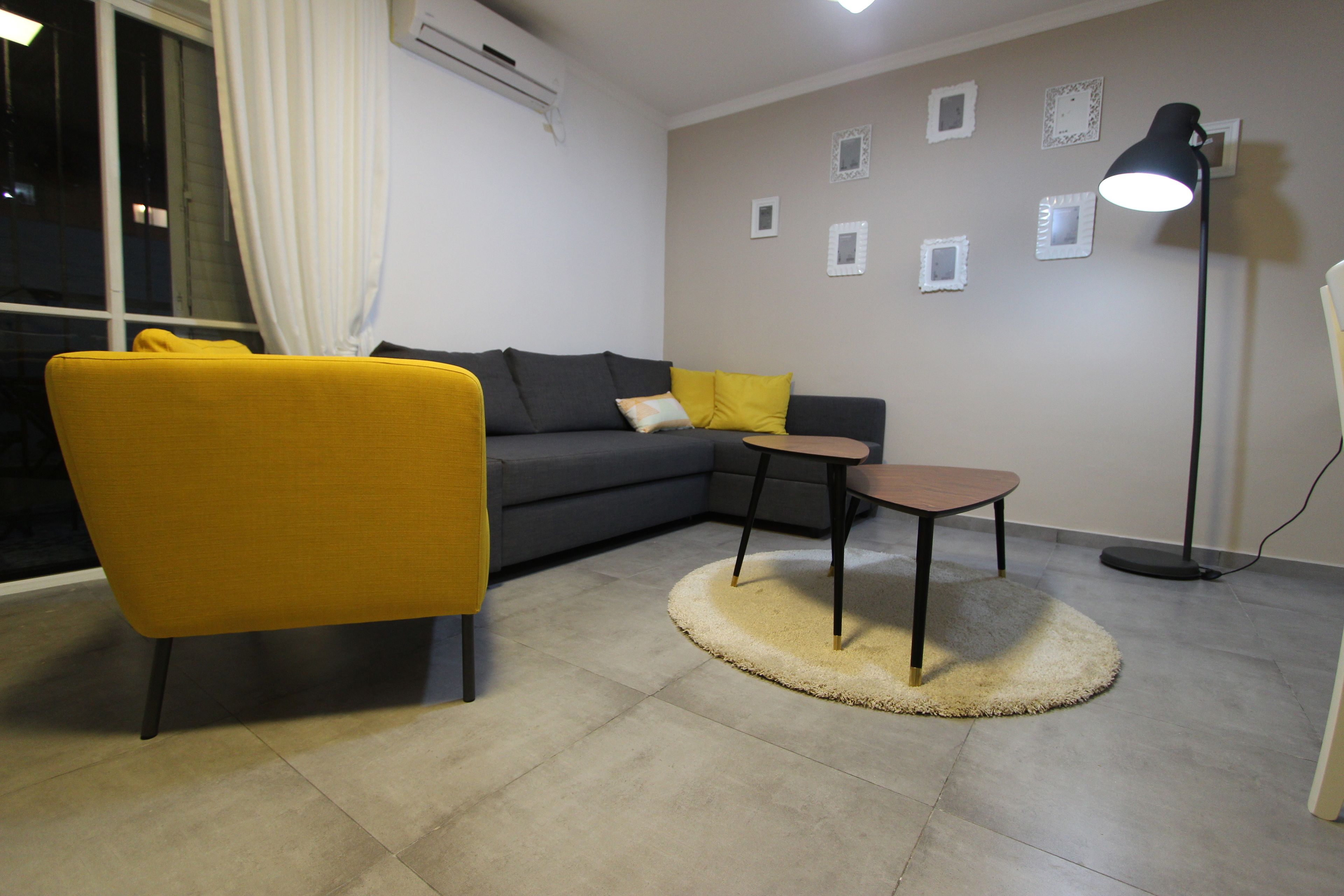 A modern living room with a yellow armchair, a grey sofa with yellow cushions, a small wooden table, and a floor lamp.