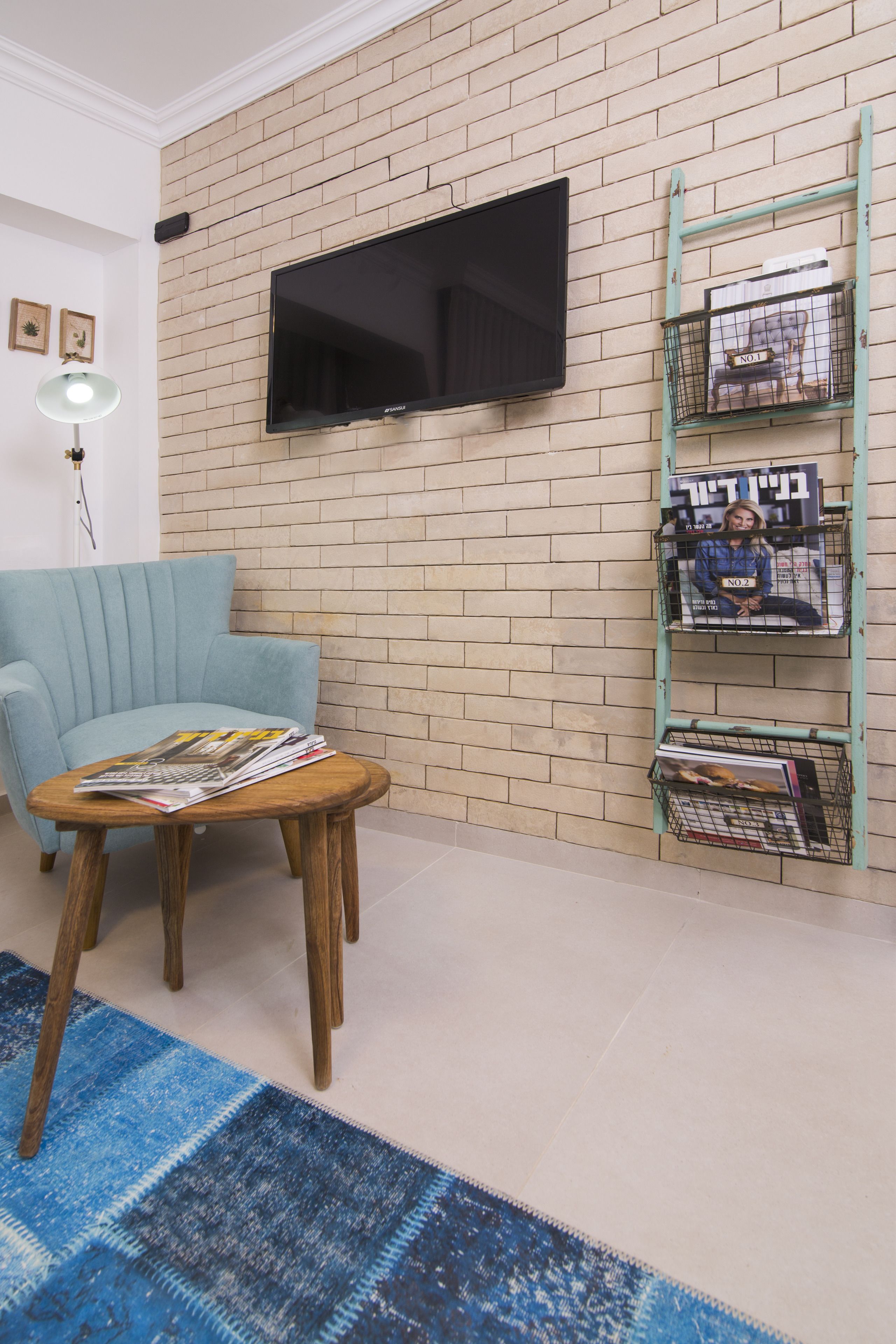 A living room with a brick wall, a mounted TV, a blue armchair, a wooden coffee table with magazines, and a blue patterned rug.