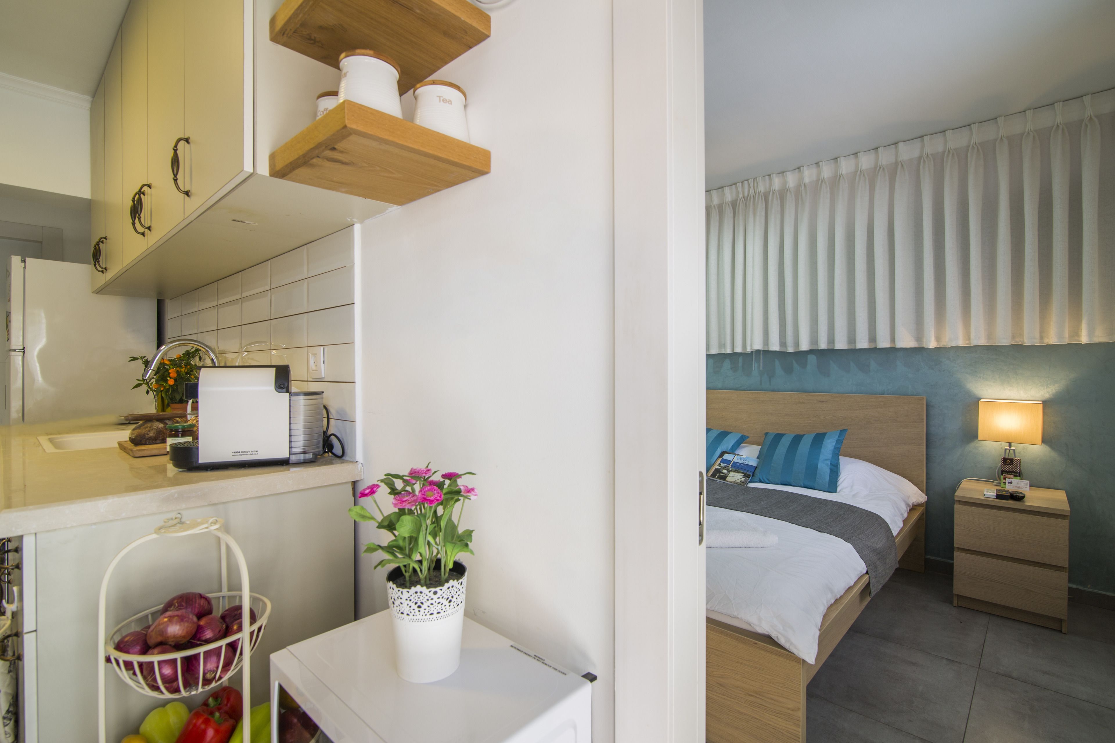 A hotel room with a bed, bedside table, and a small desk with a lamp. There is a kitchen area with a sink and cabinets.
