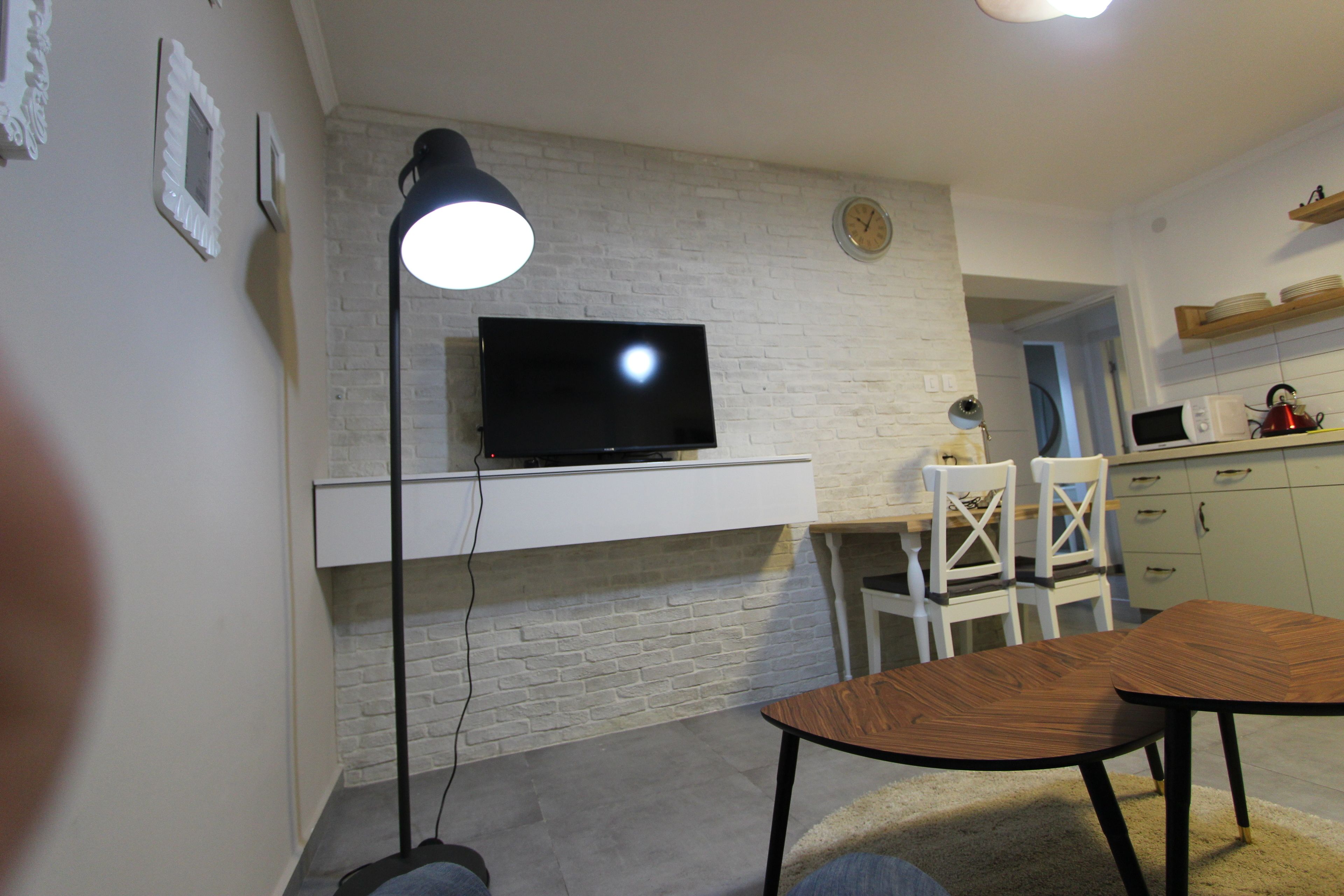 Une cuisine moderne avec des murs en briques blanches, un téléviseur à écran plat fixé au-dessus du comptoir, et un coin repas avec une table et des chaises en bois.