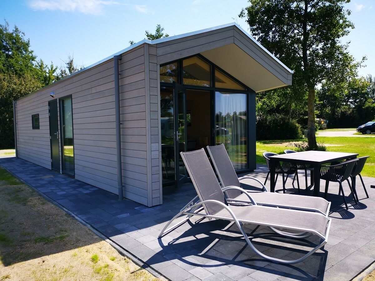 Un bâtiment moderne avec une entrée dotée d’une porte en verre, un espace extérieur aménagé avec des chaises en métal et une table, ainsi qu’une terrasse dallée.