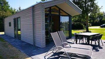 Un bâtiment moderne avec une entrée dotée d’une porte en verre, un espace extérieur aménagé avec des chaises en métal et une table, ainsi qu’une terrasse dallée.