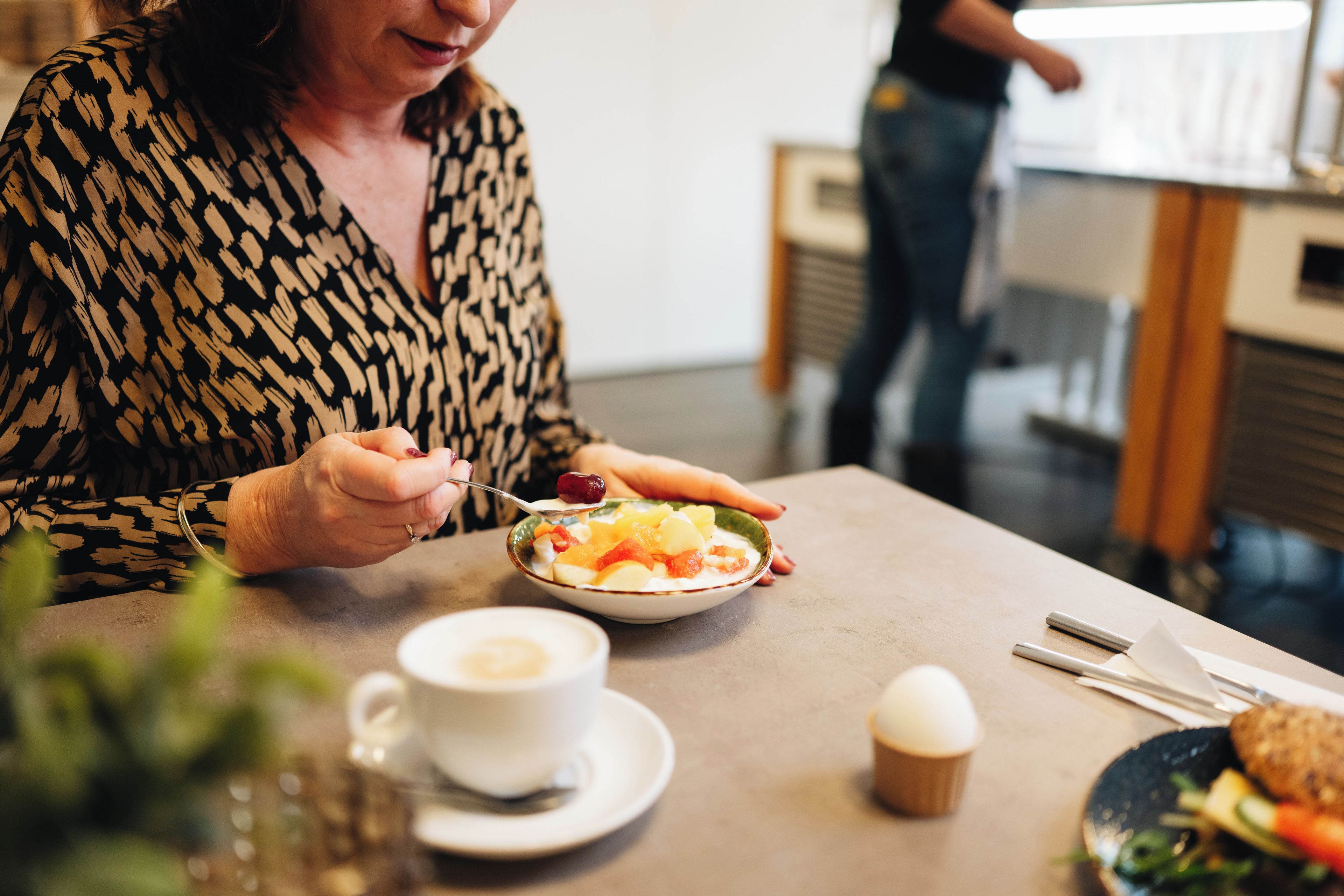 Eine Person isst an einem Tisch, auf dem eine Schüssel Essen, eine Tasse Kaffee und ein Muffin stehen.