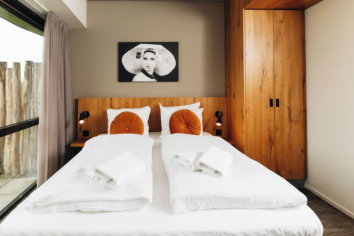 A double bed with white linens, two orange accent pillows, a wooden headboard, a wall-mounted picture, and a wooden wardrobe.