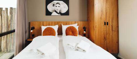 Una cama doble con sábanas blancas, dos almohadas naranjas decorativas, un cabecero de madera, un cuadro colgado en la pared y un armario de madera.