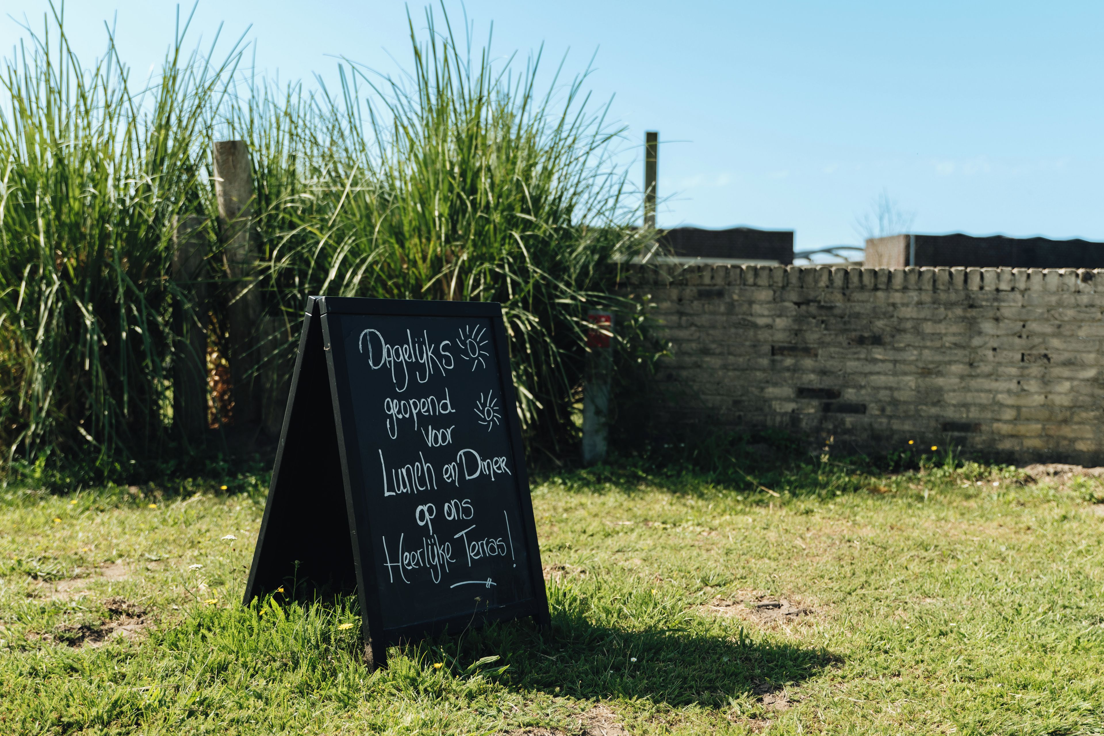 A blackboard sign with white chalk text in a grassy area with a brick wall in the background.