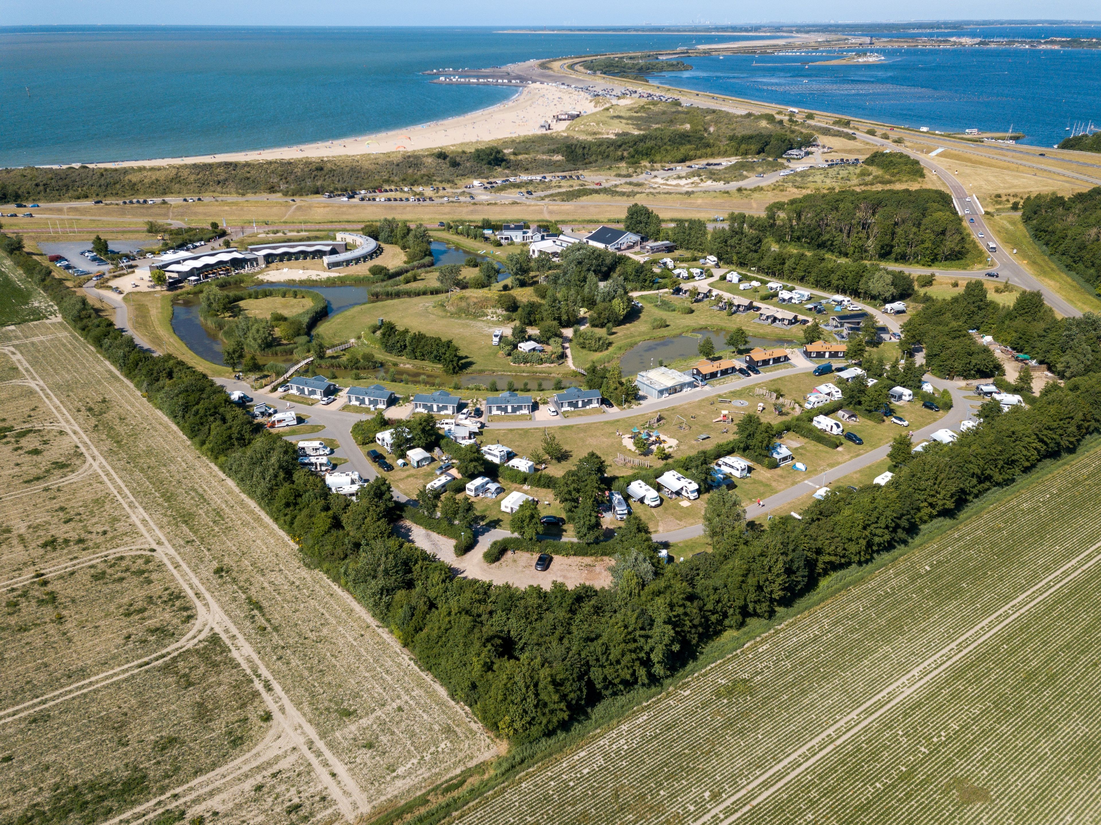 Vue aérienne d’un terrain de camping avec des camping-cars, des tentes et des bâtiments disposés autour d’un point d’eau central.