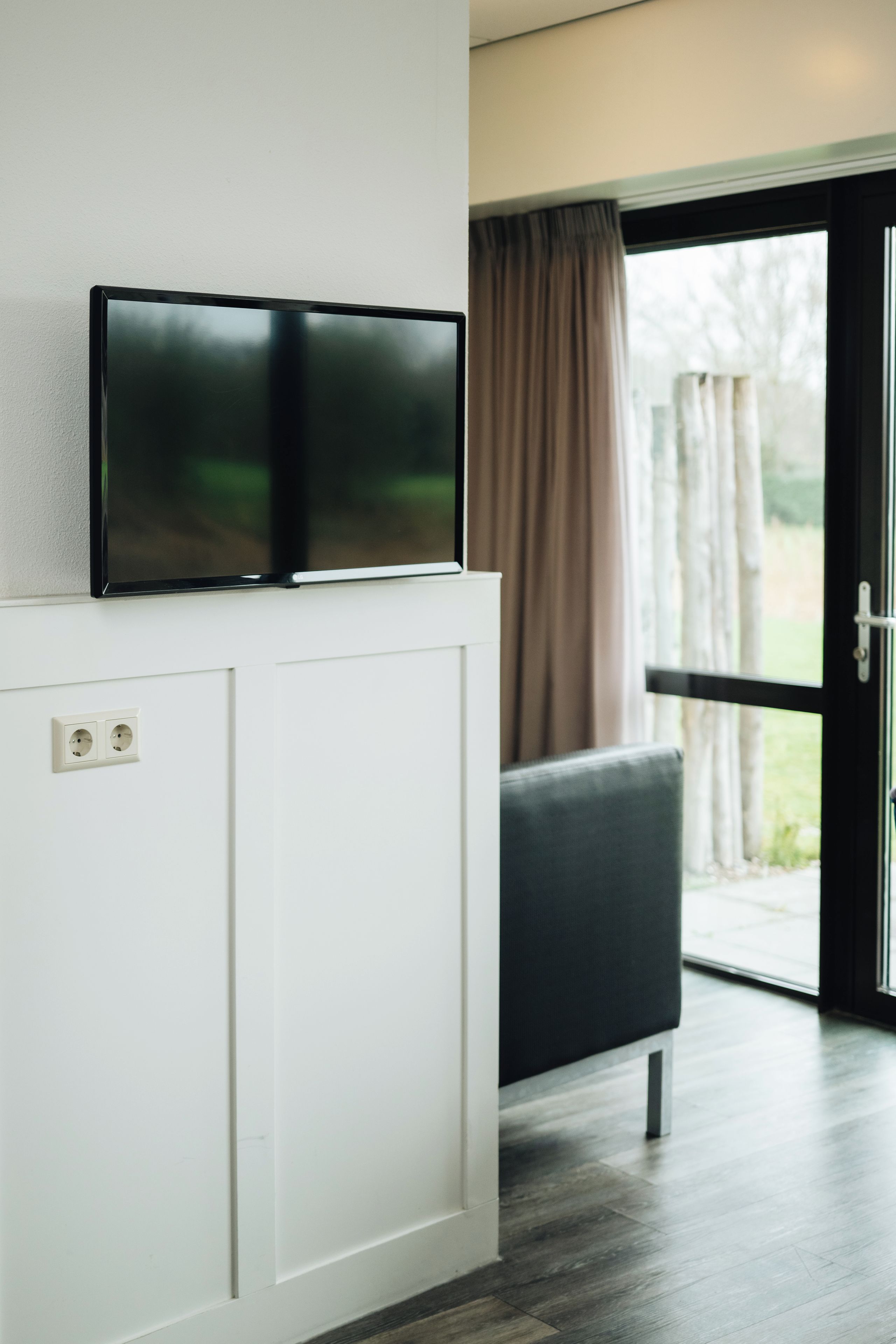 A modern room with a flat-screen TV mounted on a white panel, a black sofa, and a sliding glass door leading to an outdoor area.