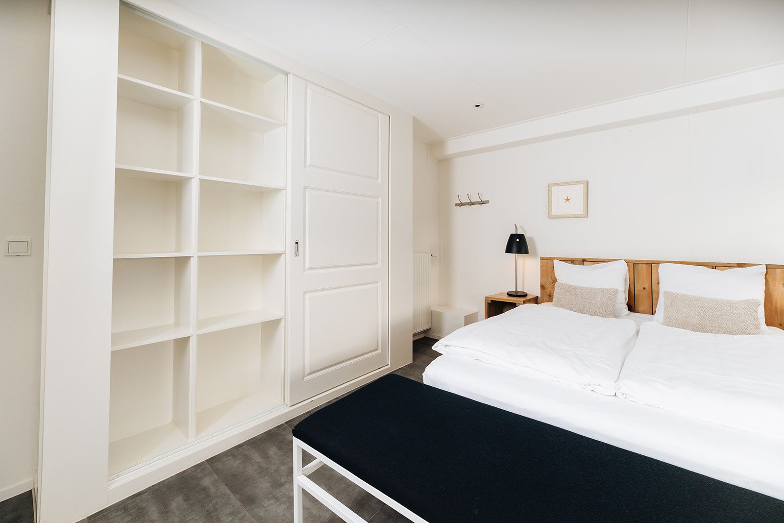 Un dormitorio con un armario blanco grande, una cama con ropa de cama blanca, una mesita de noche con una lámpara y un cuadro en la pared.