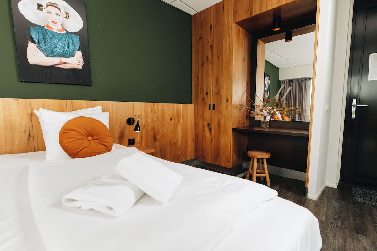 A hotel room with a bed, a wooden headboard, a painting on the wall, and a mirror reflecting a room with a window and curtains.