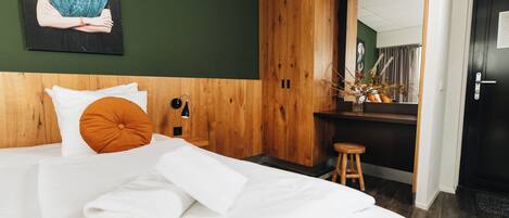 A hotel room with a bed, a wooden headboard, a painting on the wall, and a mirror reflecting a room with a window and curtains.