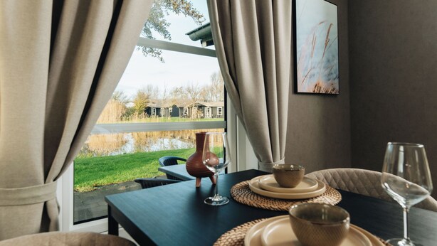 A dining table with plates, bowls, and a wine glass, set against a window with curtains, overlooking a grassy area and a building. - Resort Land en Zee (Noordwelle)