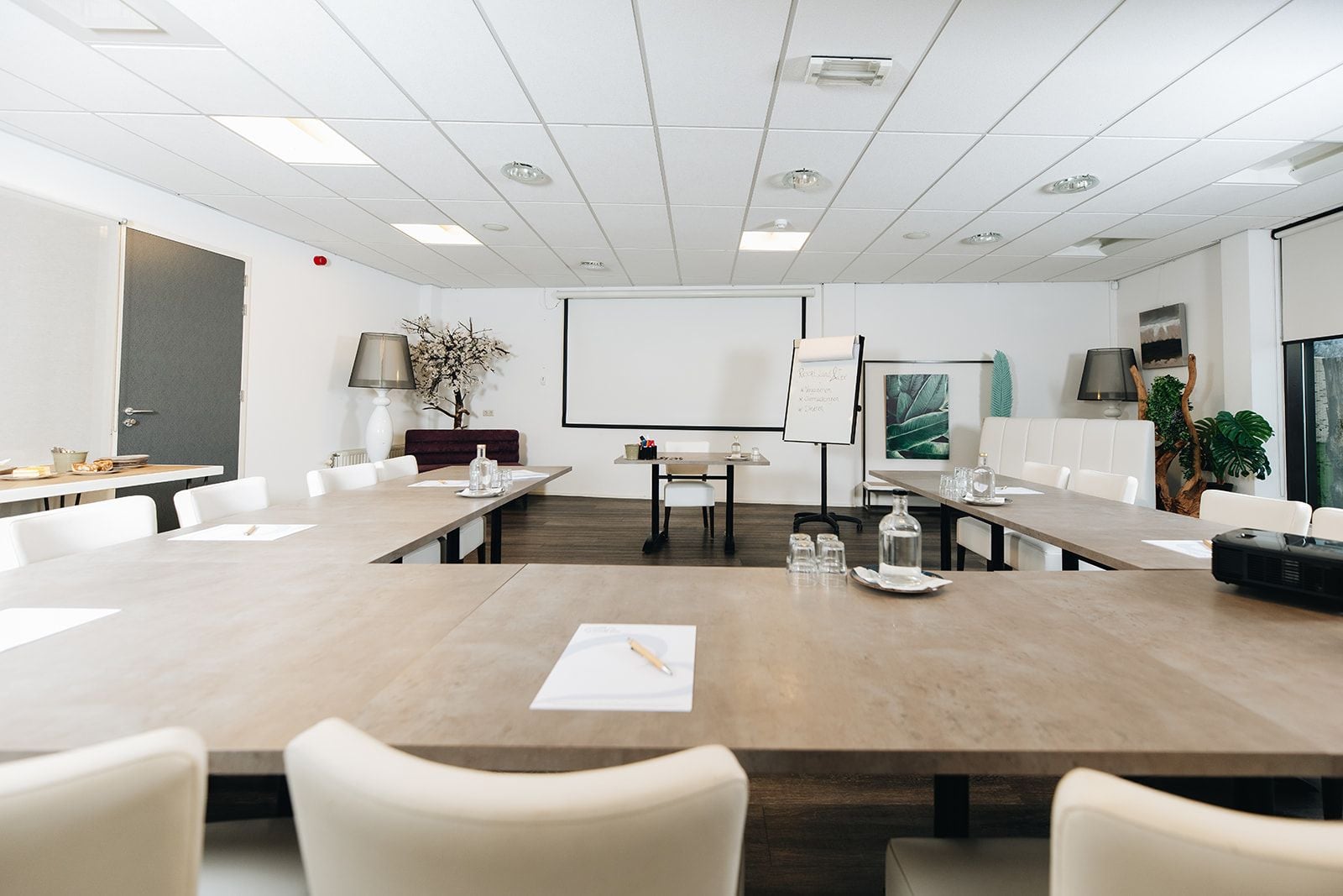 Une salle de conférence avec une longue table, des chaises blanches, un écran de projection, un tableau blanc et une plante.